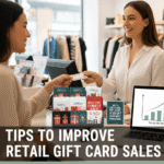 A smiling retail cashier handing a branded gift card to a customer at a checkout counter, featuring a display of various gift card designs and a laptop showing an upward-trending sales graph.